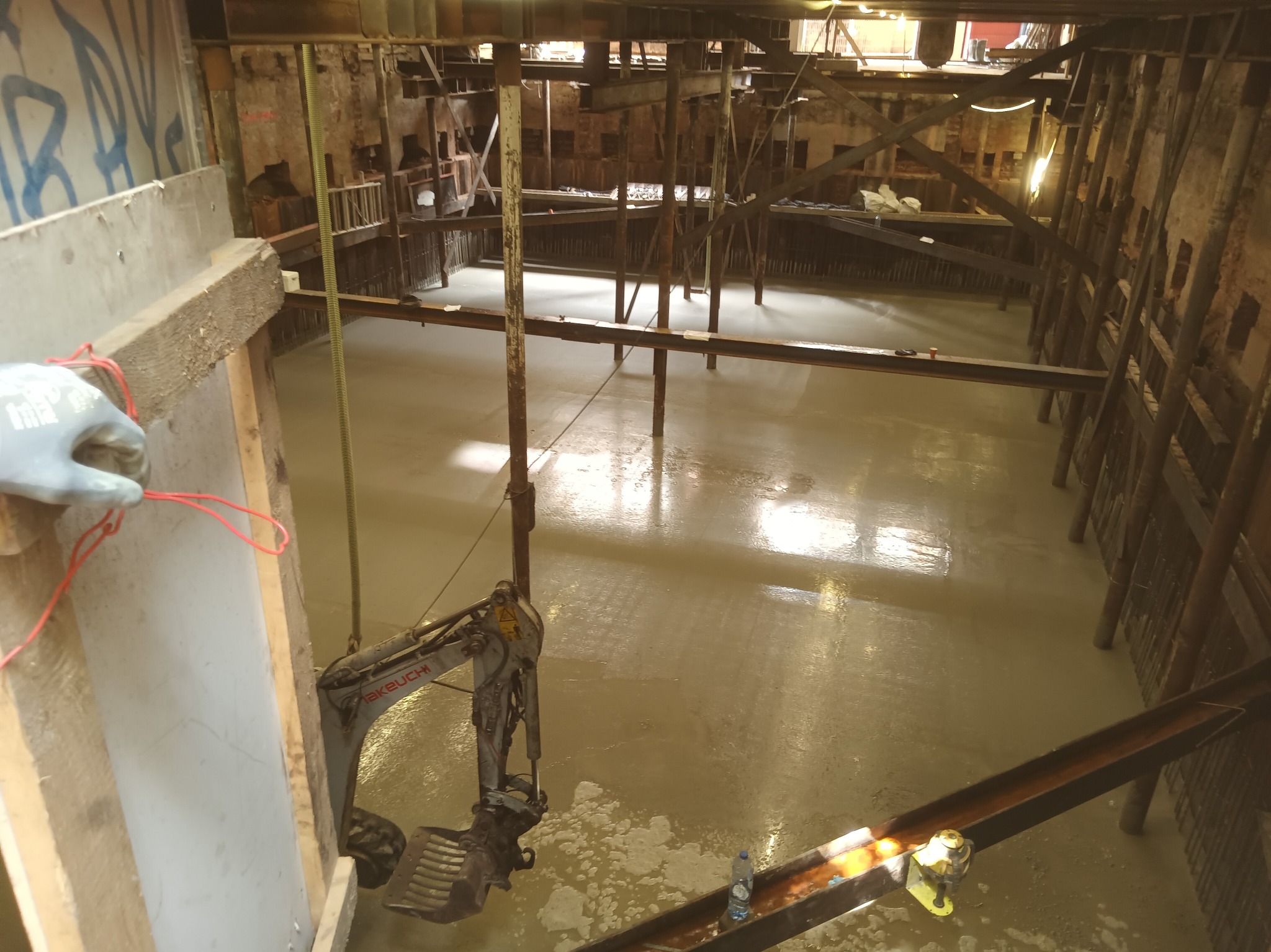 Wykop z zabezpieczeniami i wylewką betonową. Widoczny minikoparka Takeuchi, drewniane szalunki i stalowe podpory. Ujęcie z góry, ukazujące etap prac budowlanych.