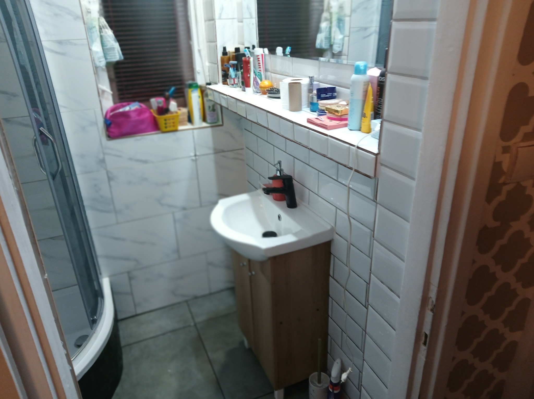 Umywalka z szafką w łazience wyłożonej białymi, prostokątnymi płytkami. W tle kabina prysznicowa i okno z żaluzjami. Na półce nad umywalką kosmetyki i akcesoria.