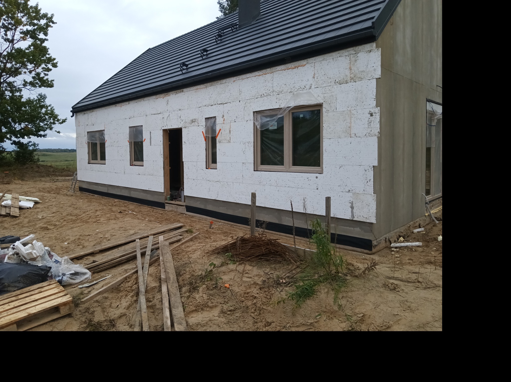 Dom w trakcie budowy: ściany z białego styropianu, czarny dach, okna z folią ochronną. Na ziemi leżą deski i materiały budowlane. Widok z zewnątrz, etap ocieplania.