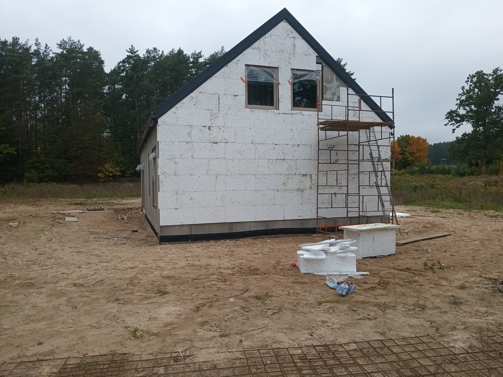 Dom w trakcie budowy z białych bloczków, widoczne rusztowanie i teren budowy. Okna zabezpieczone folią. W tle las i pochmurne niebo.