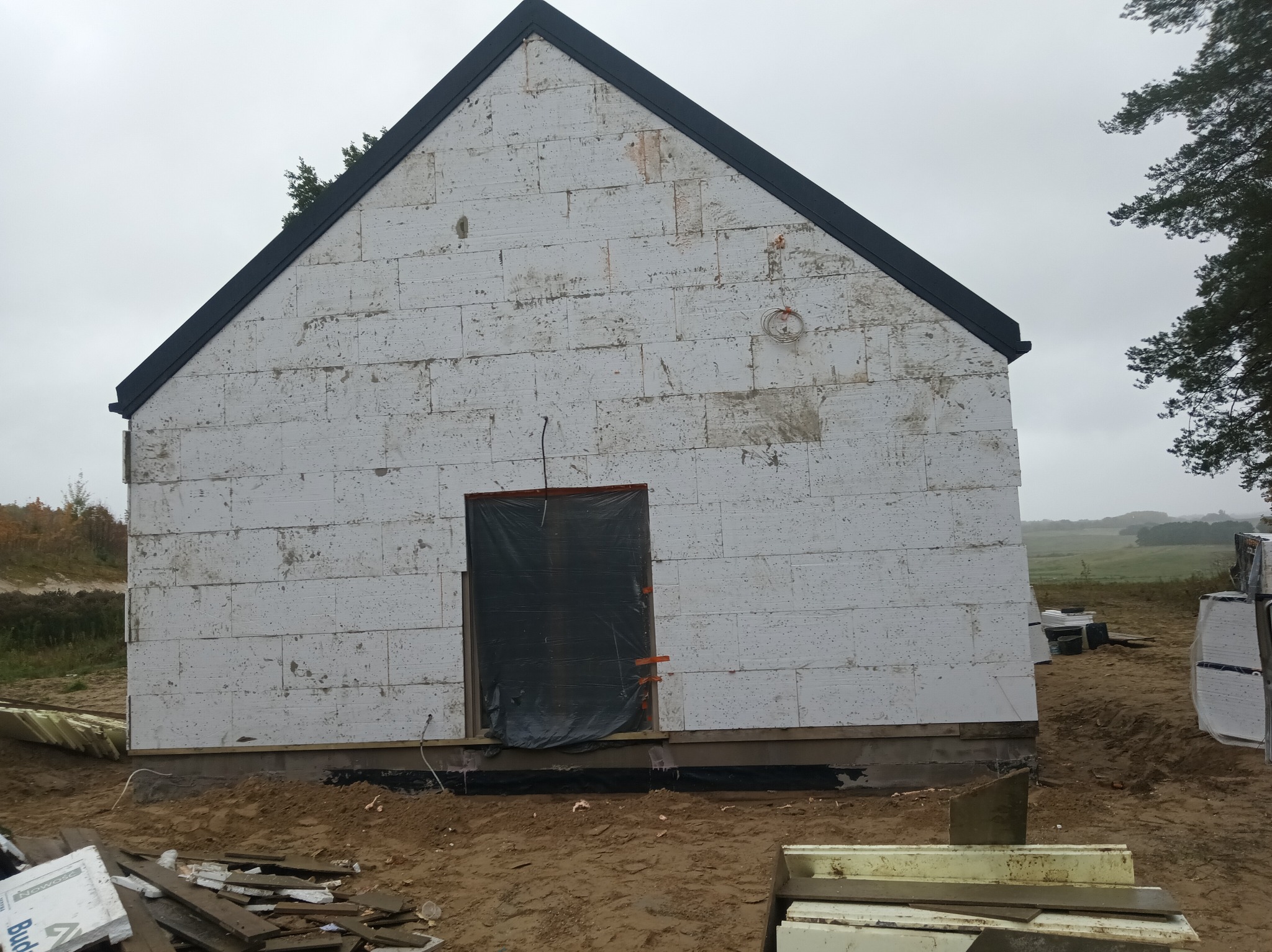 Budowa domu z białych bloczków, zabezpieczone wejście folią, widoczne materiały budowlane. Dom w trakcie budowy, z czarnym dachem i widocznym krajobrazem w tle.