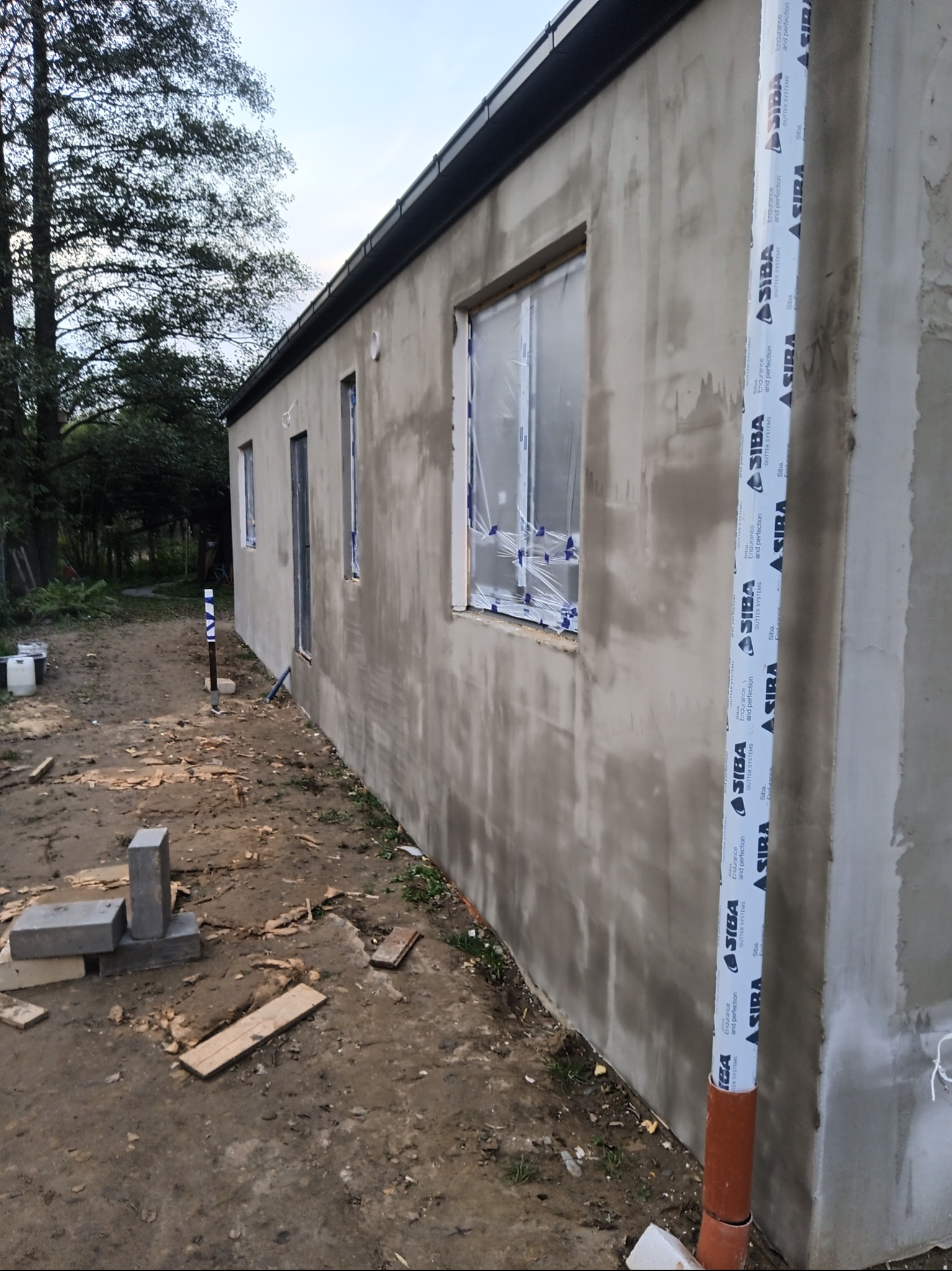 Surowy budynek po nałożeniu szarego tynku zewnętrznego, z oknami zabezpieczonymi folią i rurą spustową z logo SIBA, na tle ziemi i drzew.