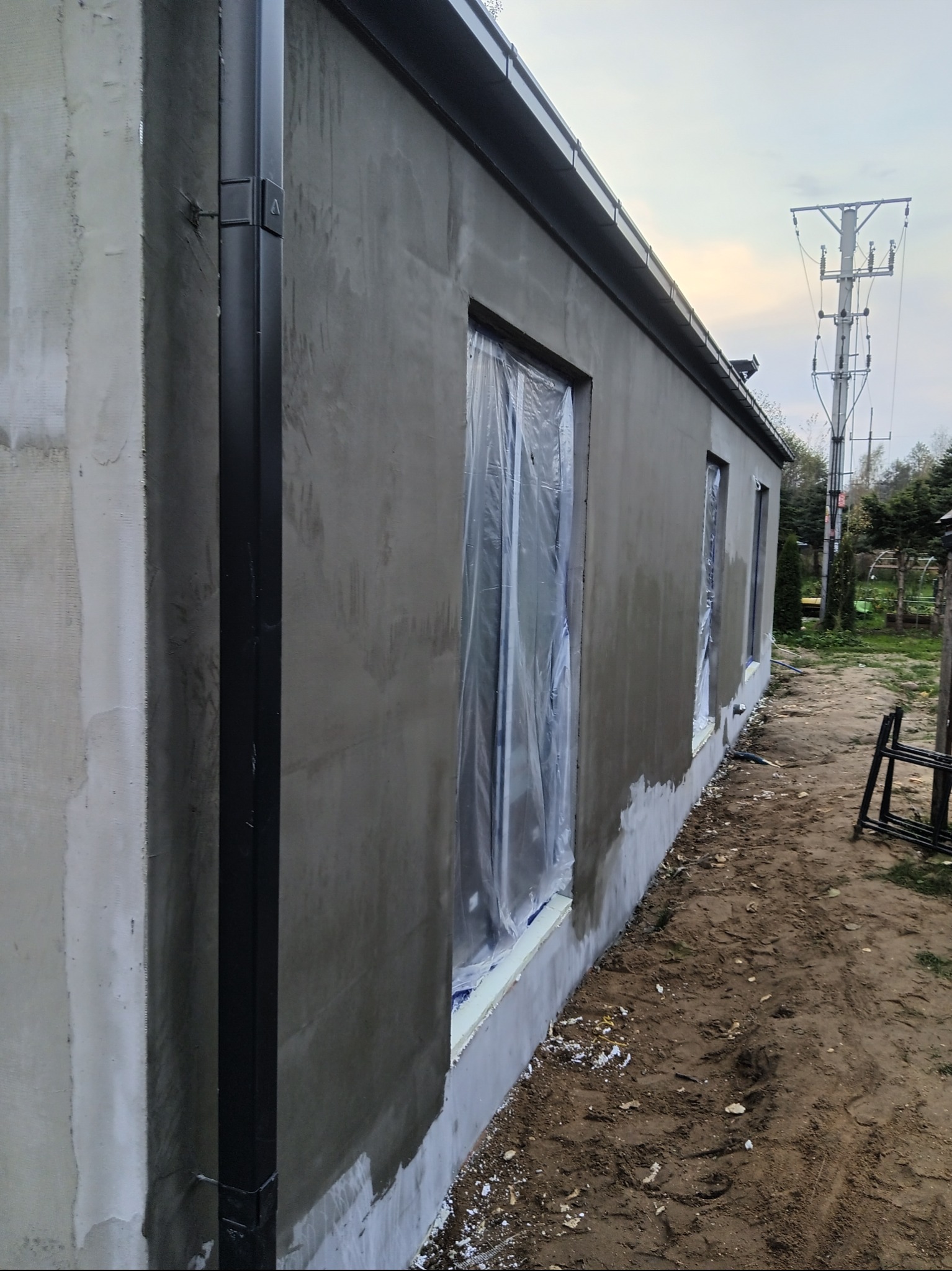 Ściana budynku w trakcie prac wykończeniowych: świeżo nałożony tynk, okna zabezpieczone folią, widoczny system rynnowy. Ujęcie z perspektywy ziemi.