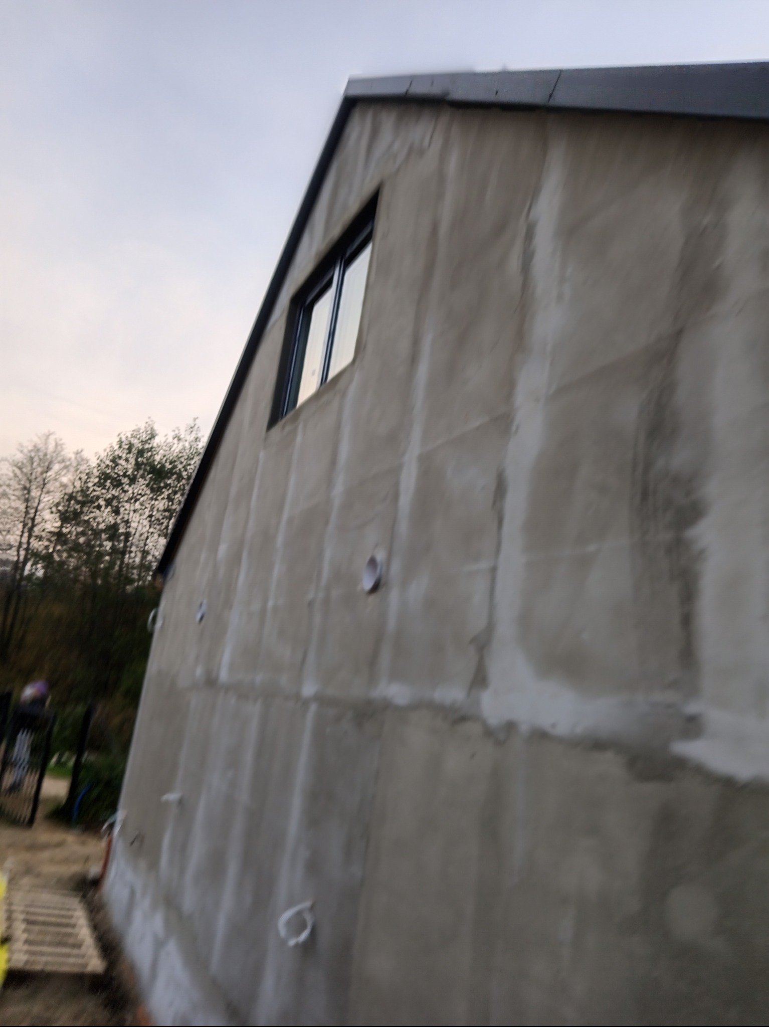 Surowa ściana budynku z oknem, w trakcie prac wykończeniowych. Widoczne niedoskonałości tynku i elementy instalacji. Tło stanowią drzewa i niebo.