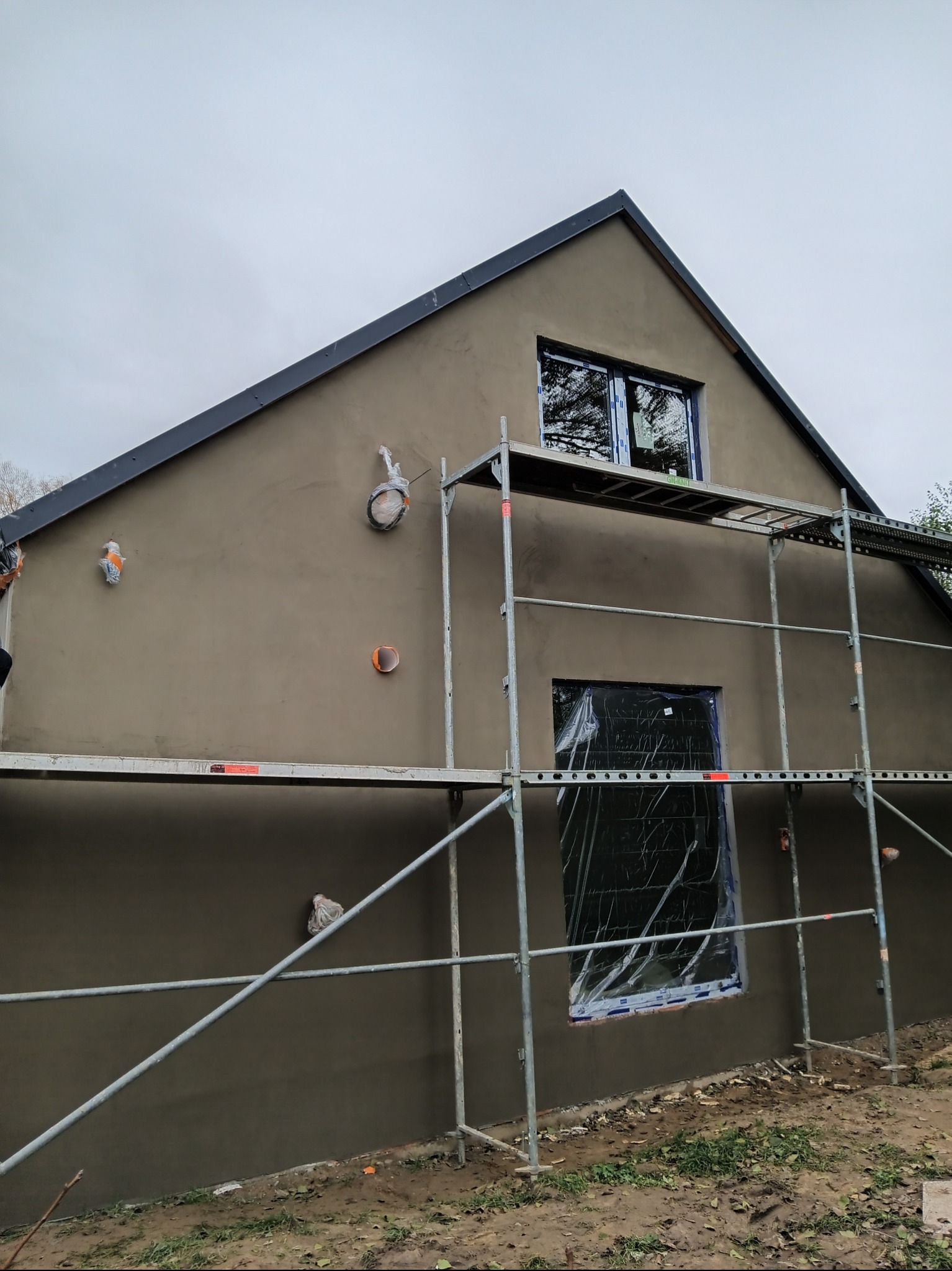 Elewacja domu w trakcie prac wykończeniowych: świeżo nałożony tynk w odcieniach szarości, okna zabezpieczone folią, widoczne rusztowania. Detale instalacji na ścianie.
