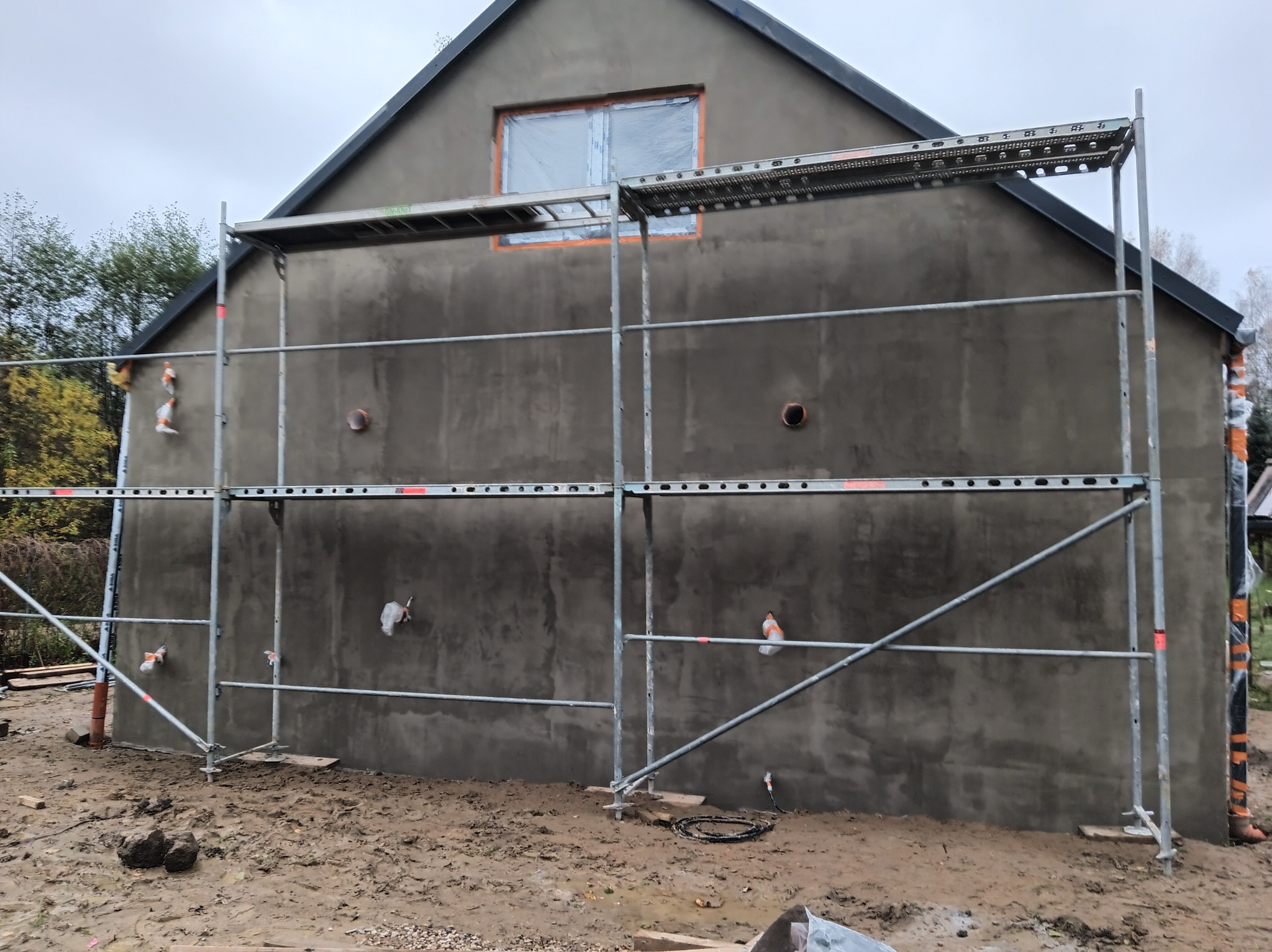 Świeżo otynkowana ściana domu jednorodzinnego z widocznymi rusztowaniami i zabezpieczonym oknem, ukazująca etap prac wykończeniowych na zewnątrz budynku.