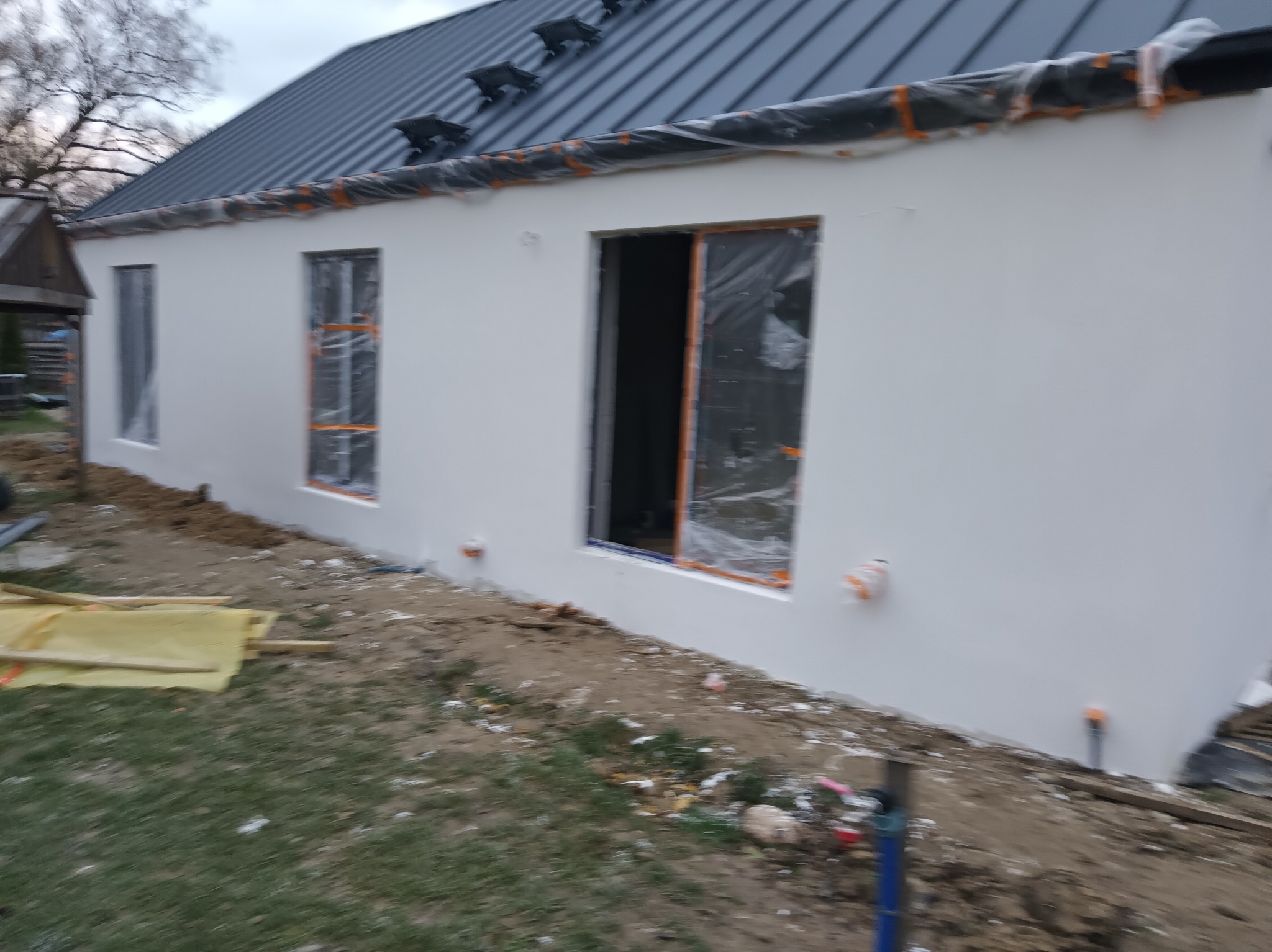 Biała elewacja domu z szarą blachodachówką w trakcie prac wykończeniowych. Okna i drzwi zabezpieczone folią, widoczne elementy instalacji na zewnątrz.