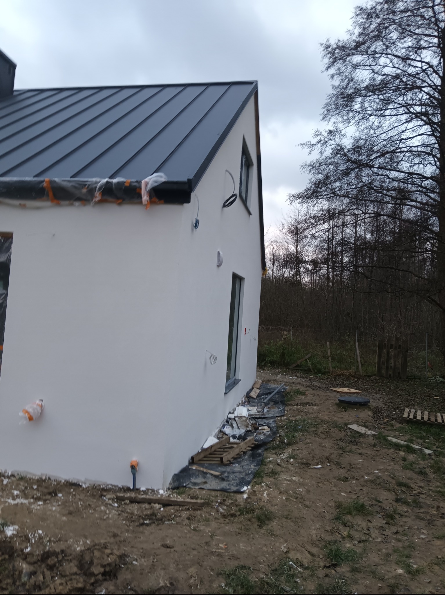 Boczny widok na świeżo otynkowany, biały dom z ciemnym dachem na tle pochmurnego nieba i drzew. Widoczne elementy instalacji i zabezpieczenia okien folią.