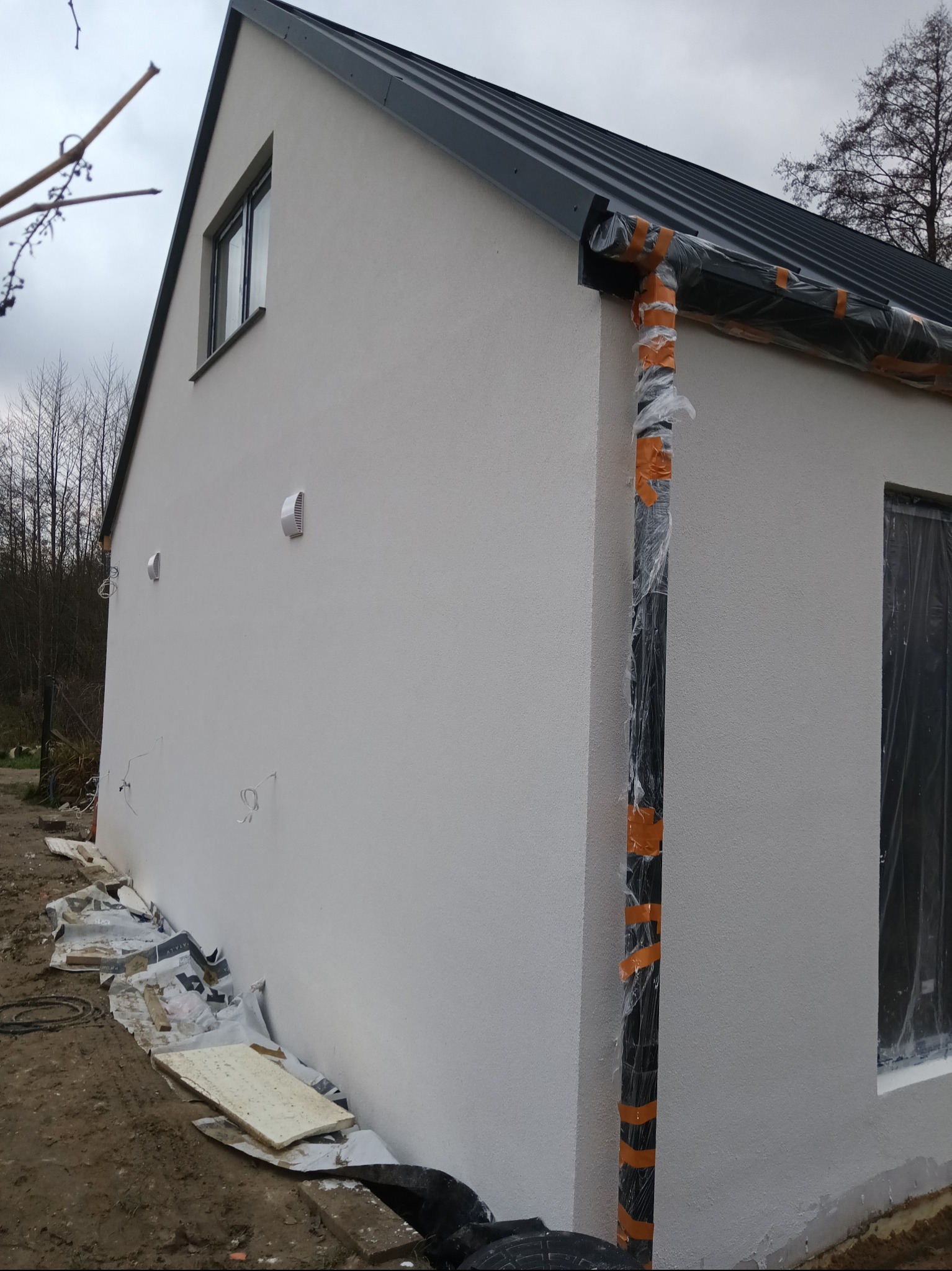 Elewacja budynku po nałożeniu tynku strukturalnego, z zabezpieczonymi elementami (rynna, okna) folią i taśmą. Widoczne fragmenty instalacji i resztki materiałów budowlanych.