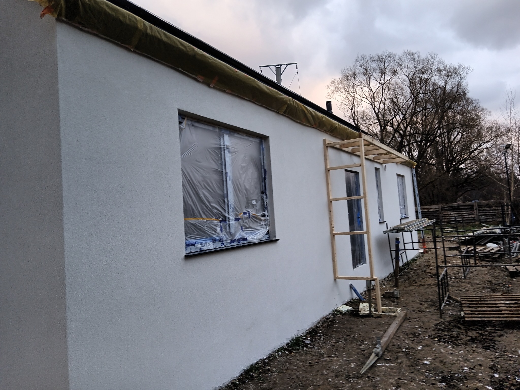 Ocieplony budynek z nałożonym tynkiem, okna zabezpieczone folią, widoczne rusztowanie i materiały budowlane. Prace wykończeniowe na zewnątrz budynku.