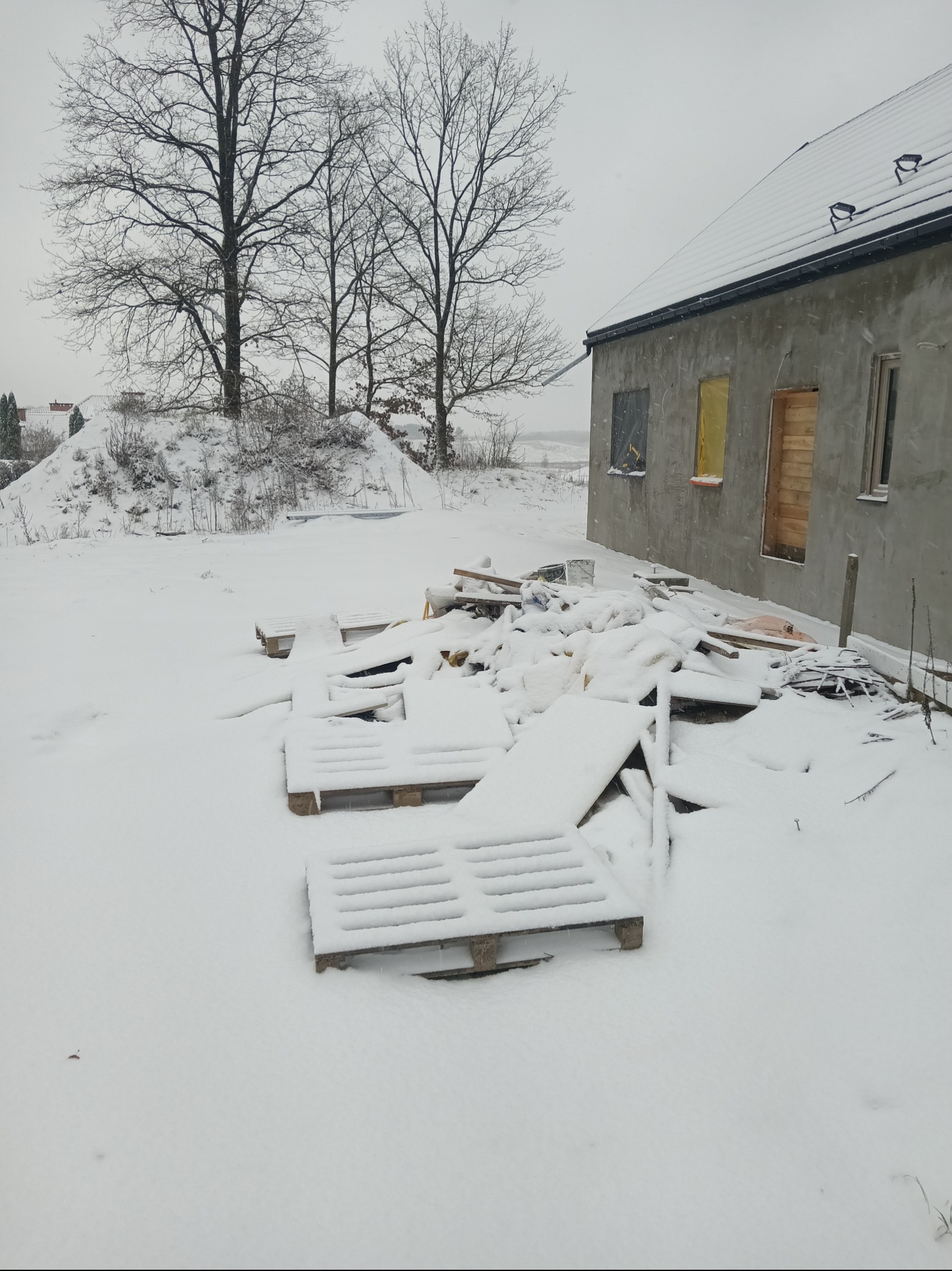 Surowy budynek w zimowej scenerii: śnieg pokrywa teren, palety i materiały budowlane. Okna zabite deskami, drzewa bez liści w tle. Szary, pochmurny dzień.