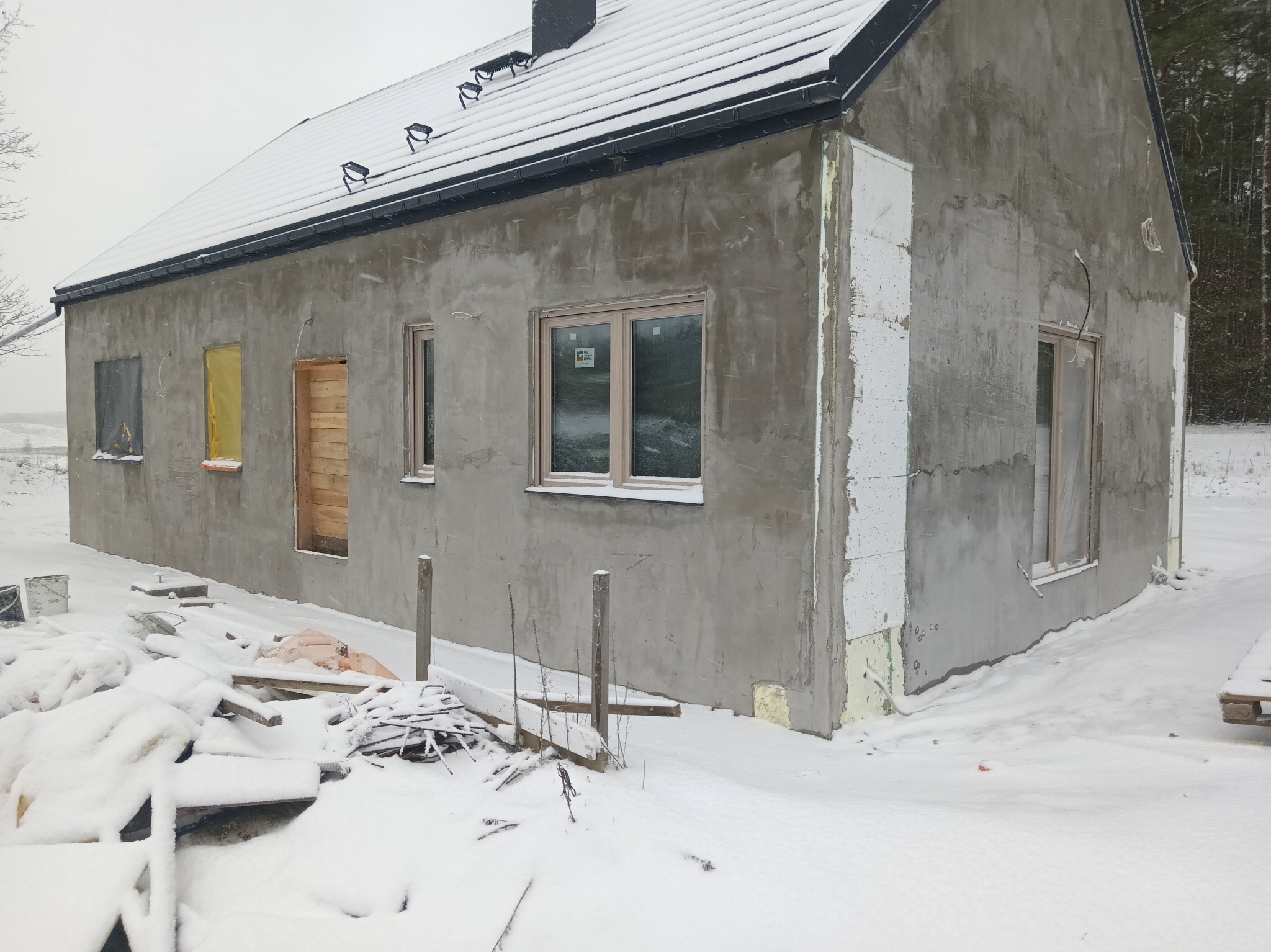 Surowy dom jednorodzinny w zimowej scenerii, z oknami i drzwiami w trakcie montażu, ocieplony częściowo styropianem, pokryty szarym tynkiem. Śnieg na dachu i wokół budynku.