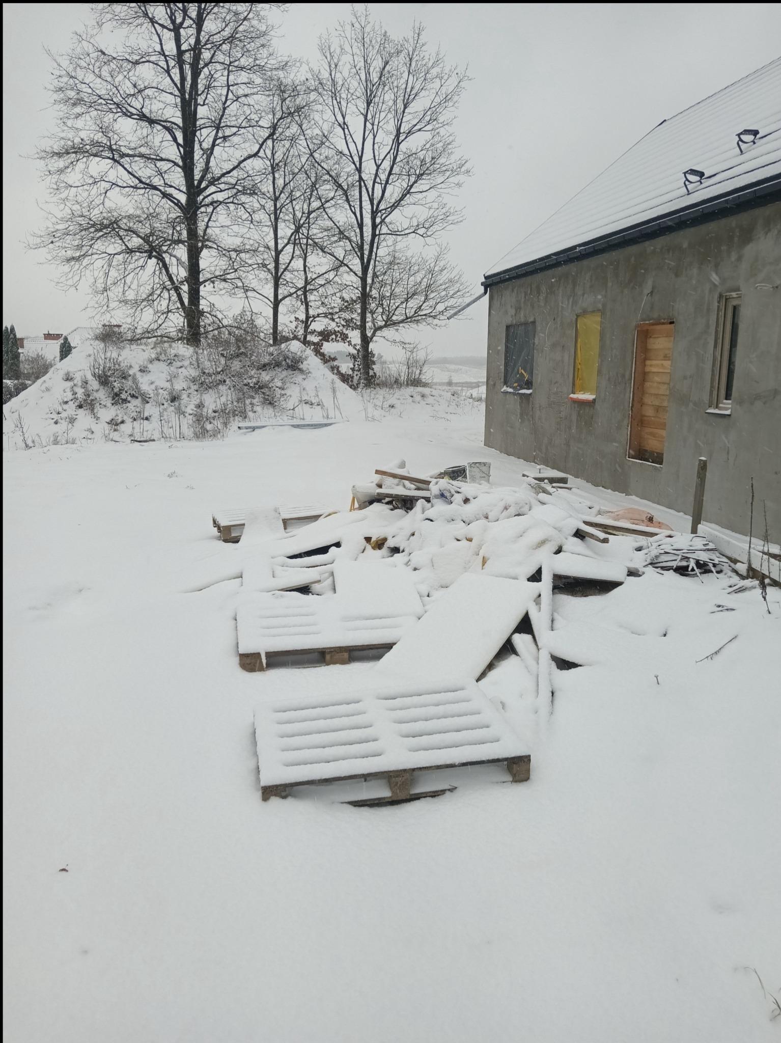 Surowy budynek w zimowej scenerii, z oknami zabezpieczonymi folią i deskami, otoczony śniegiem i porozrzucanymi materiałami budowlanymi. Widoczne drzewa bez liści.