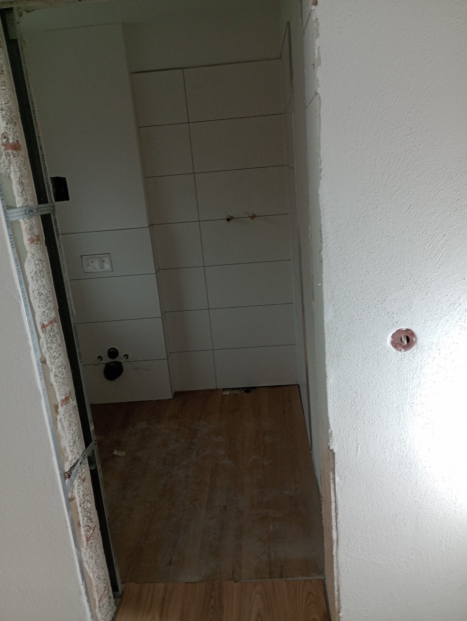 Wnętrze łazienki w trakcie remontu: widoczne białe płytki na ścianach, drewnopodobna podłoga i metalowa konstrukcja przy ścianie. Otwór w ścianie z instalacją.