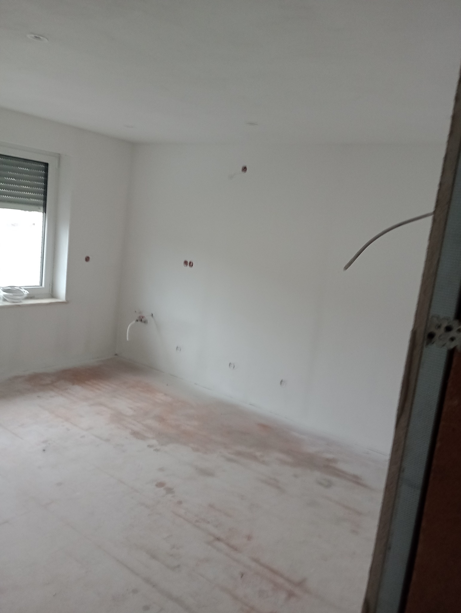 Wnętrze pokoju w trakcie prac wykończeniowych: białe ściany z widocznymi instalacjami elektrycznymi i hydraulicznymi, surowa podłoga, okno z roletą. Widoczny fragment futryny.