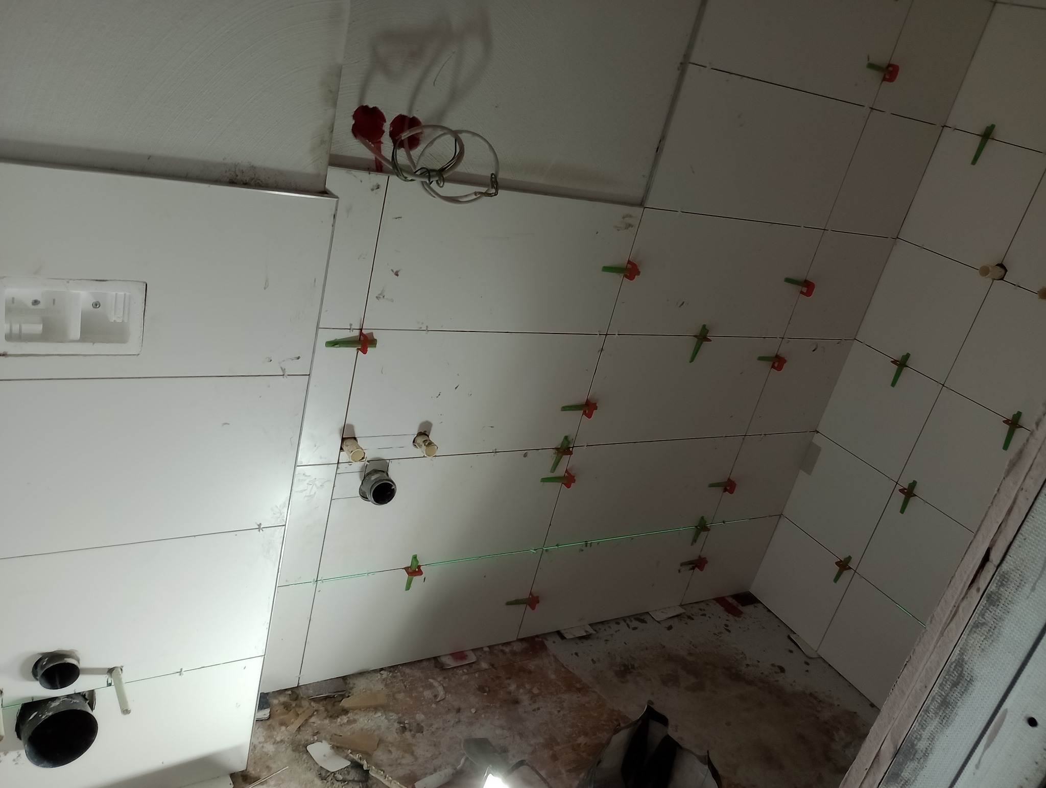 Białe płytki ścienne w trakcie montażu, z użyciem poziomicy laserowej i klipsów poziomujących. Widoczne instalacje hydrauliczne i fragment podłogi w stanie surowym.