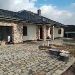 Nowo wybudowany parterowy dom z ceglaną elewacją i wybrukowanym podjazdem z kostki, widoczne pojemniki budowlane i materiały wykończeniowe.