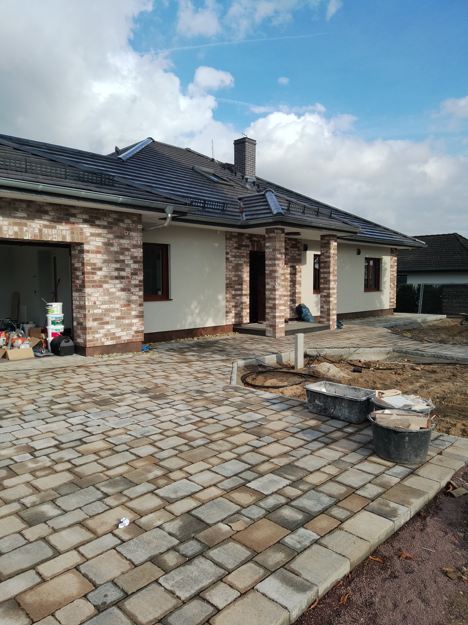 Nowo wybudowany parterowy dom z ceglaną elewacją i wybrukowanym podjazdem z kostki, widoczne pojemniki budowlane i materiały wykończeniowe.