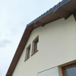 Fragment jasnej elewacji domu z drewnianą podbitką dachową w kolorze brązowym i oknami z drewnianymi ramami, widok z dołu na tle pochmurnego nieba.