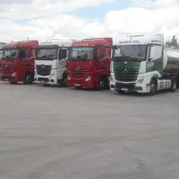 Rząd zaparkowanych ciężarówek różnych marek (Nanek-Pol, Vectura) na betonowym placu przed halą magazynową w pochmurny dzień.