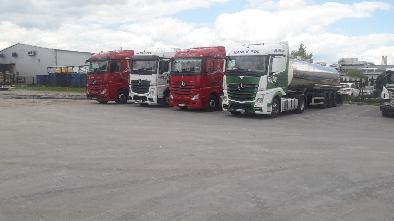 Rząd zaparkowanych ciężarówek różnych marek (Nanek-Pol, Vectura) na betonowym placu przed halą magazynową w pochmurny dzień.