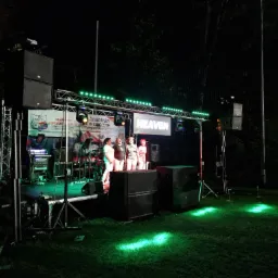 Zespół muzyczny na scenie z oświetleniem podczas koncertu plenerowego. Widoczne nagłośnienie, baner z nazwą zespołu 'Heaven' i publiczność.