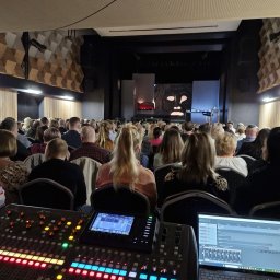 NEXA ENERGY Leszek Korycki - Konsoleta dźwiękowa z laptopem na tle widowni w sali widowiskowej z dekoracją w stylu etno na scenie. Oświetlenie sceniczne włączone.