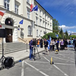 NEXA ENERGY Leszek Korycki - Ekipa filmowa rejestruje wydarzenie przed budynkiem z flagami Polski i UE. Widoczny kamerzysta, mikrofony i grupa osób w tle. Dzień słoneczny.