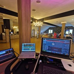 NEXA ENERGY Leszek Korycki - Sprzęt DJ'ski (mikser, laptopy) rozstawiony w sali bankietowej, gotowy do obsługi imprezy. Widoczni goście w tle, oświetlenie dekoracyjne.
