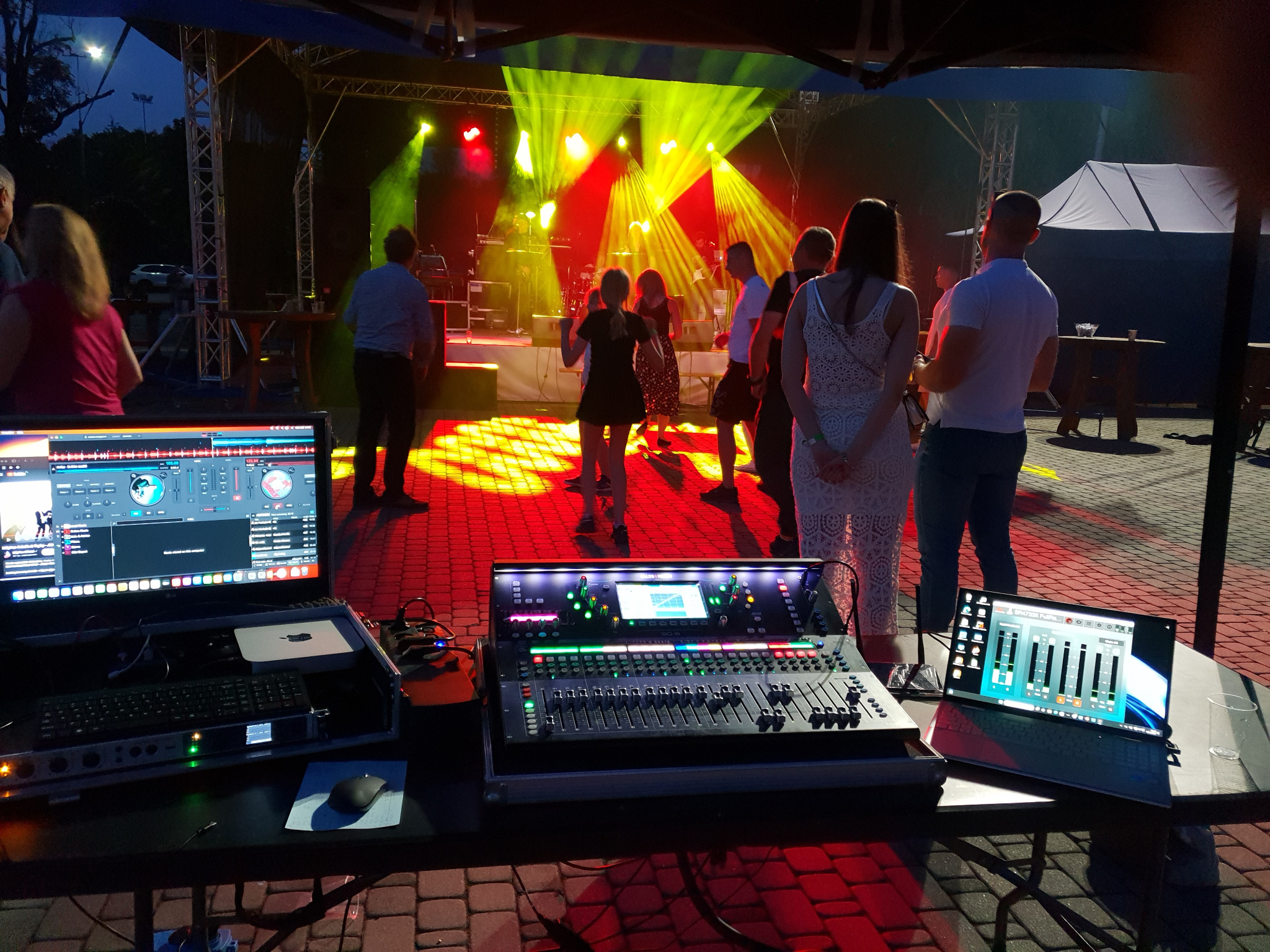 Koncert plenerowy z oświetleniem scenicznym, nagłośnieniem i tańczącymi osobami. Widoczny mikser dźwięku, laptopy i oświetlenie estradowe w kolorach czerwieni i zieleni.