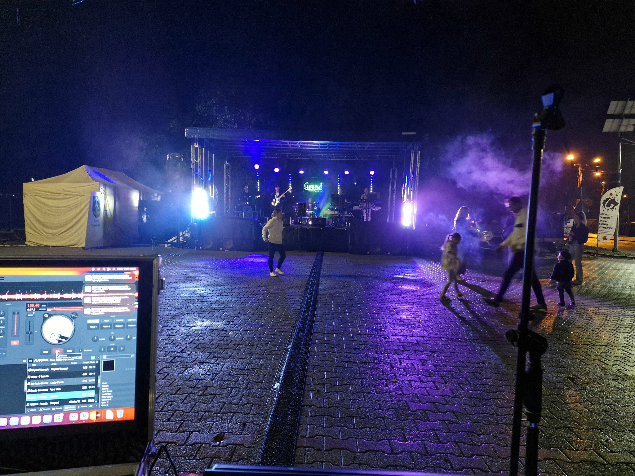 Zespół muzyczny na scenie podczas wieczornego eventu, widok zza konsolety DJ-a, z efektem mgły i oświetleniem. W tle widoczni ludzie.