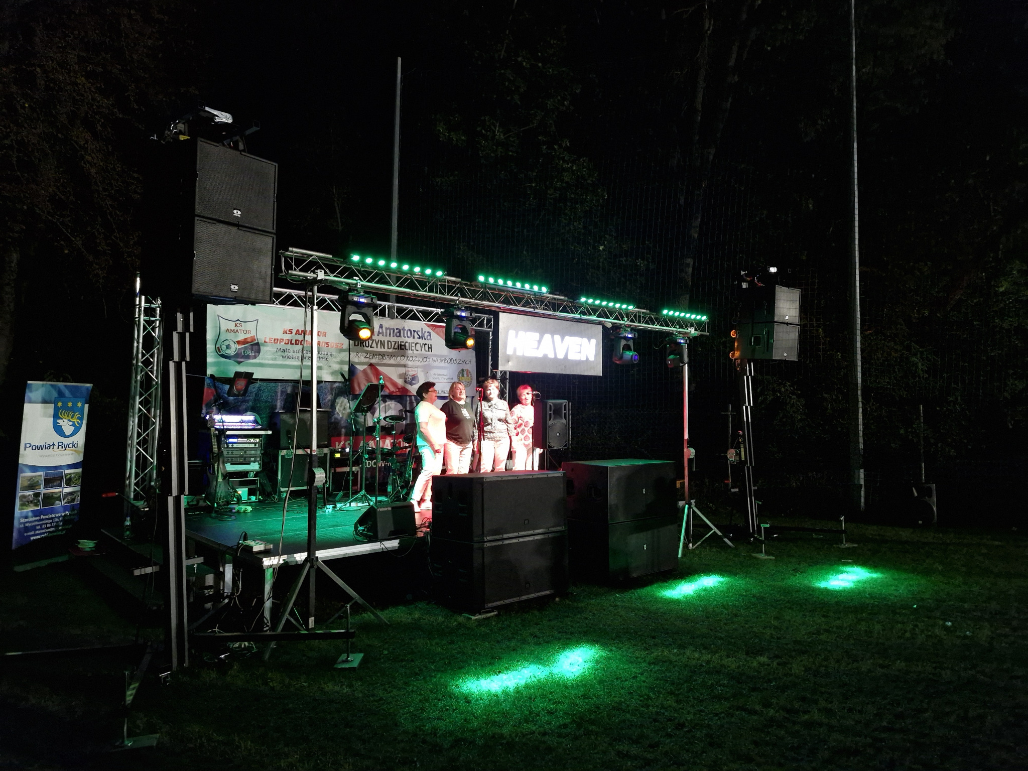 Zespół muzyczny na scenie z oświetleniem podczas koncertu plenerowego. Widoczne nagłośnienie, baner z nazwą zespołu 'Heaven' i publiczność.