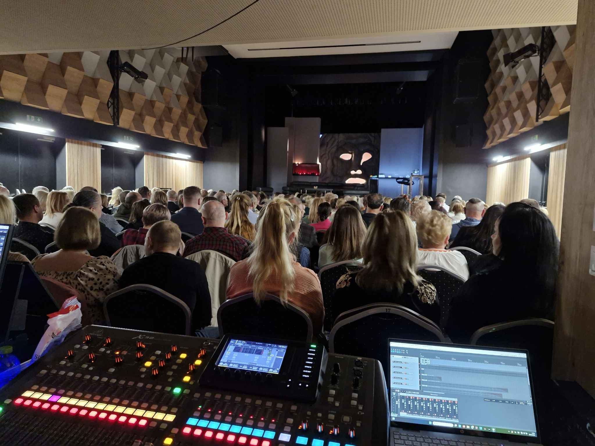 Konsoleta dźwiękowa z laptopem na tle widowni w sali widowiskowej z dekoracją w stylu etno na scenie. Oświetlenie sceniczne włączone.