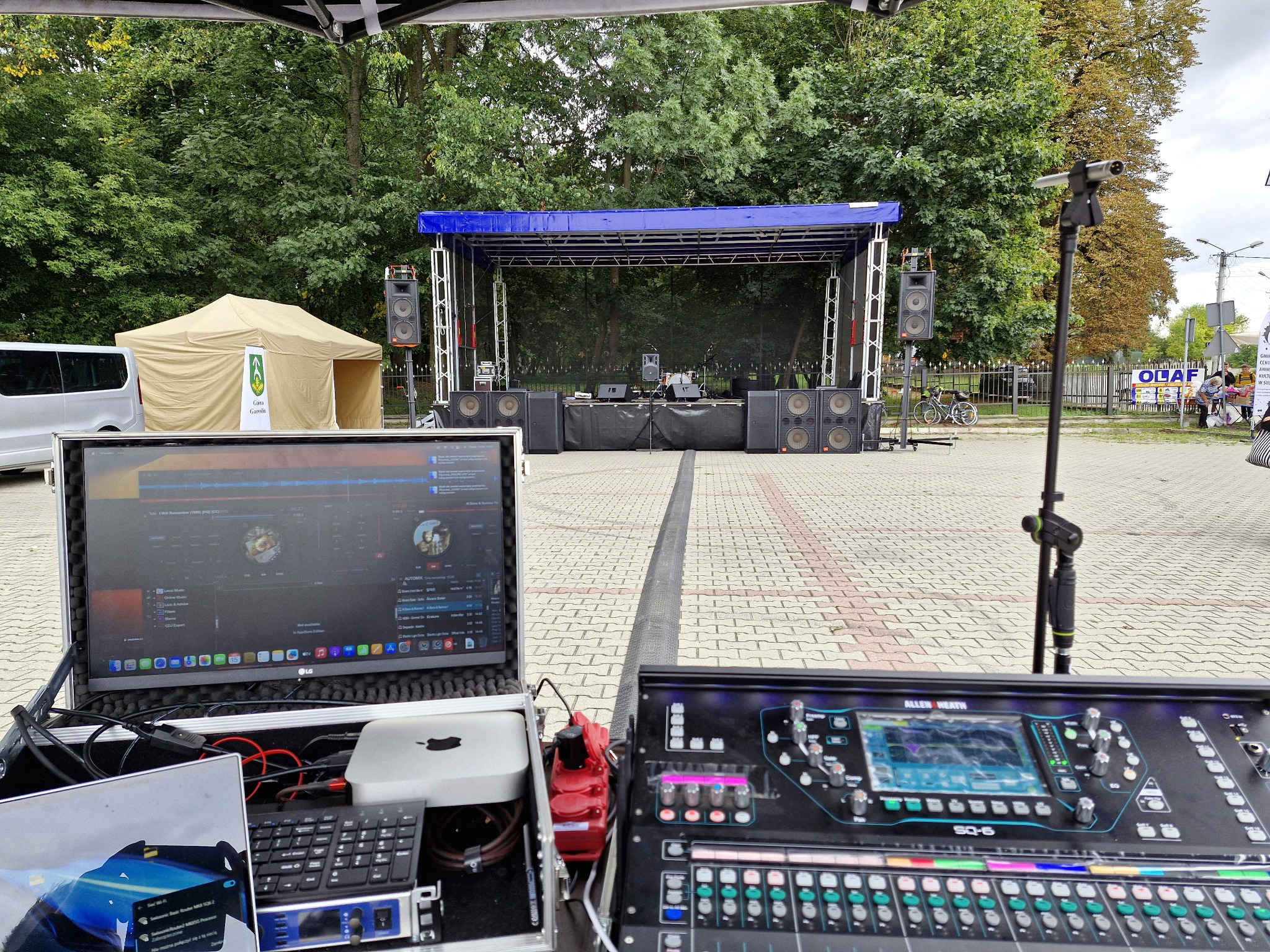 Scena plenerowa z nagłośnieniem i sprzętem DJ-skim. Widoczny mikser Allen&Heath SQ-6, laptop LG z oprogramowaniem muzycznym. W tle drzewa i namiot imprezowy.