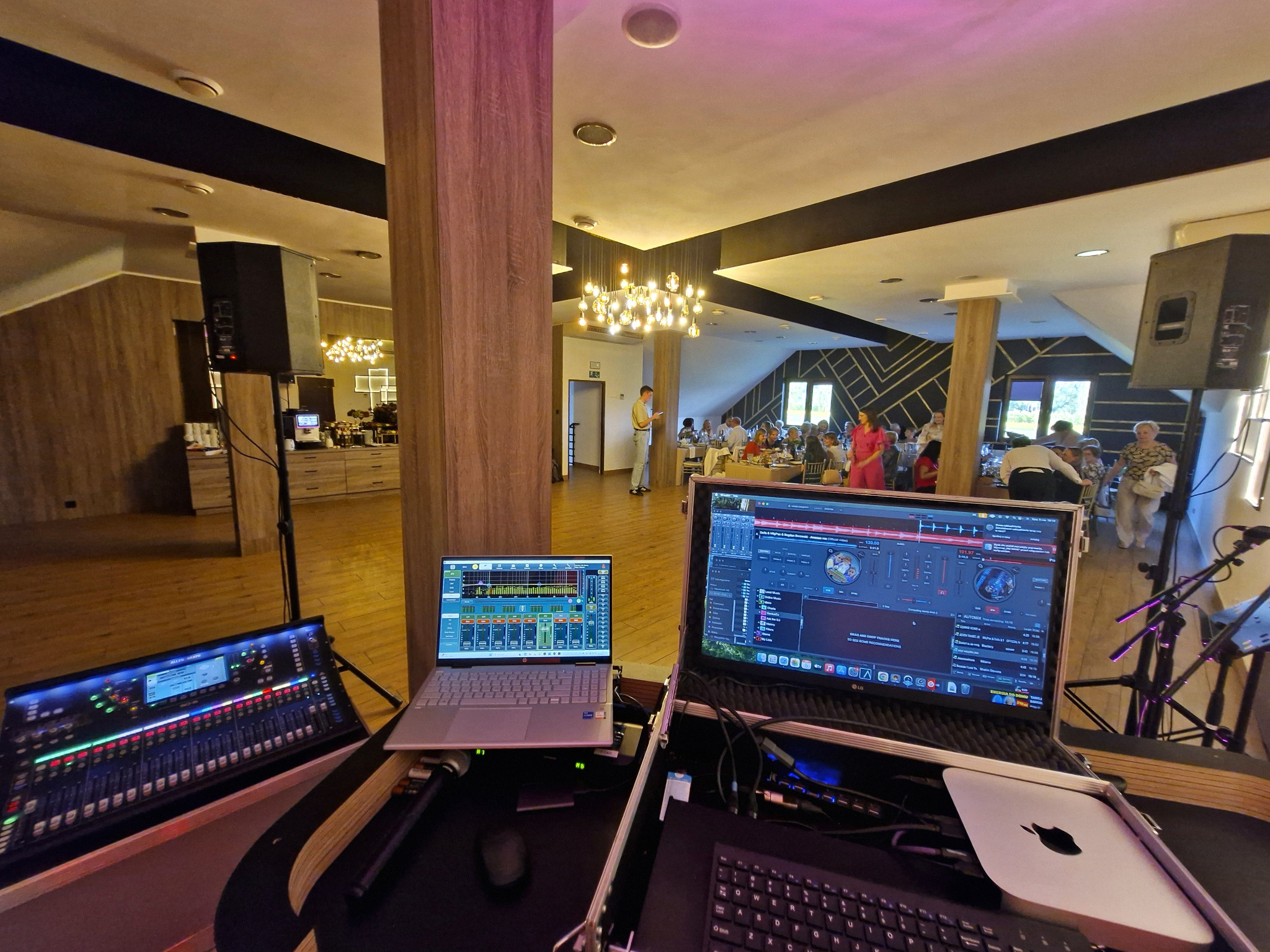 Sprzęt DJ'ski (mikser, laptopy) rozstawiony w sali bankietowej, gotowy do obsługi imprezy. Widoczni goście w tle, oświetlenie dekoracyjne.