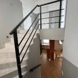 Wnętrze domu w trakcie wykańczania: schody wyłożone płytkami z czarną, metalową balustradą z poziomymi szczeblami, widok na podłogę z parkietem w jodełkę i fragment ceglanej kolumny.