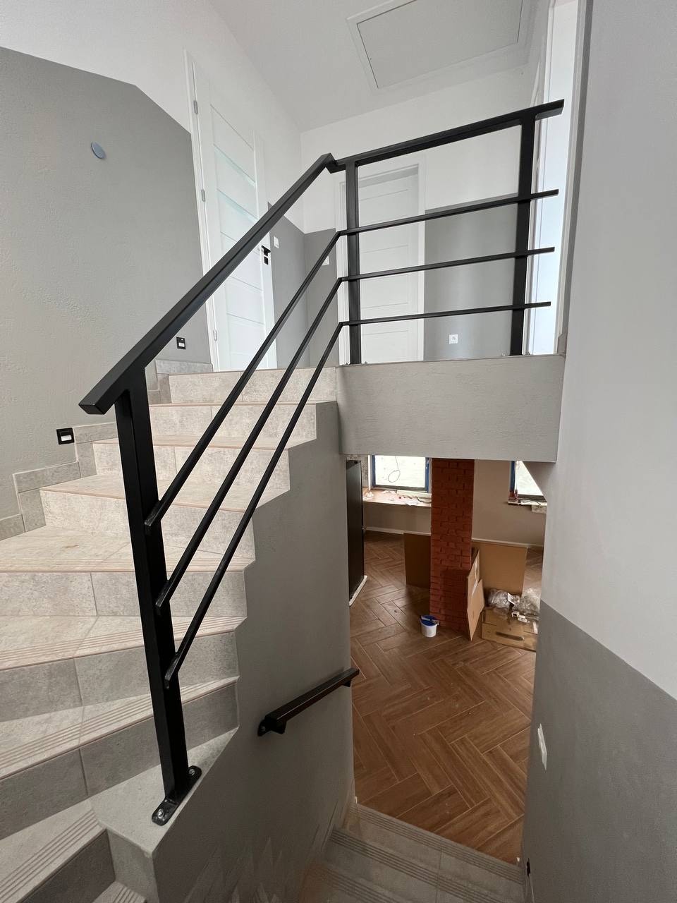 Wnętrze domu w trakcie wykańczania: schody wyłożone płytkami z czarną, metalową balustradą z poziomymi szczeblami, widok na podłogę z parkietem w jodełkę i fragment ceglanej kolumny.