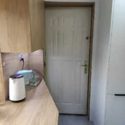 Fragment kuchni z blatem z płyty drewnopodobnej, na którym stoi biały czajnik elektryczny i dzbanek filtrujący wodę; w tle białe drzwi.