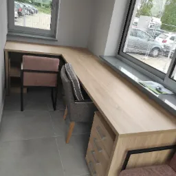 Narożne biurko z blatem imitującym jasne drewno, dopasowane do dwóch okien, z szarą komodą i dwoma krzesłami w odcieniach szarości i różu.