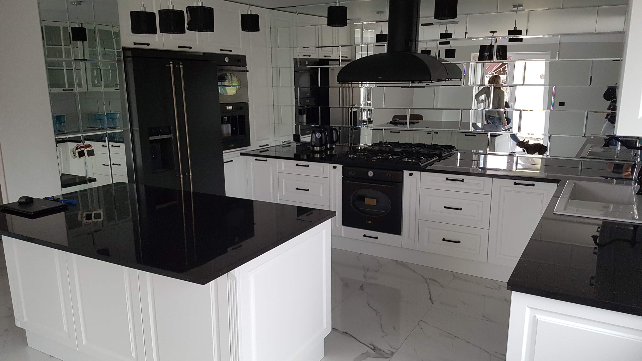 Nowoczesna kuchnia z białymi szafkami i czarnym blatem, lustrzana ściana z odbiciami, czarne lampy wiszące i marmurowa podłoga.