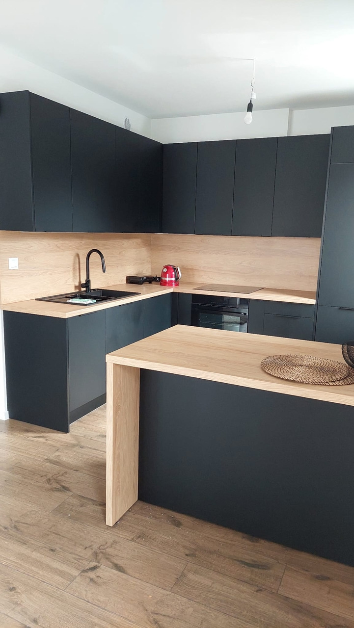 Nowoczesna kuchnia z czarnymi szafkami i blatem z jasnego drewna, widok na czarny zlew, płytę indukcyjną i czerwony czajnik na blacie.