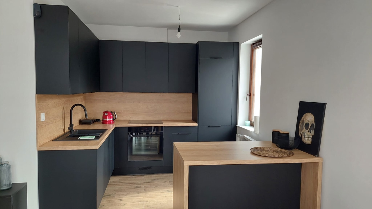 Nowoczesna kuchnia z czarnymi szafkami i drewnianymi blatami, czerwony czajnik na blacie, obraz z czaszką w tle.