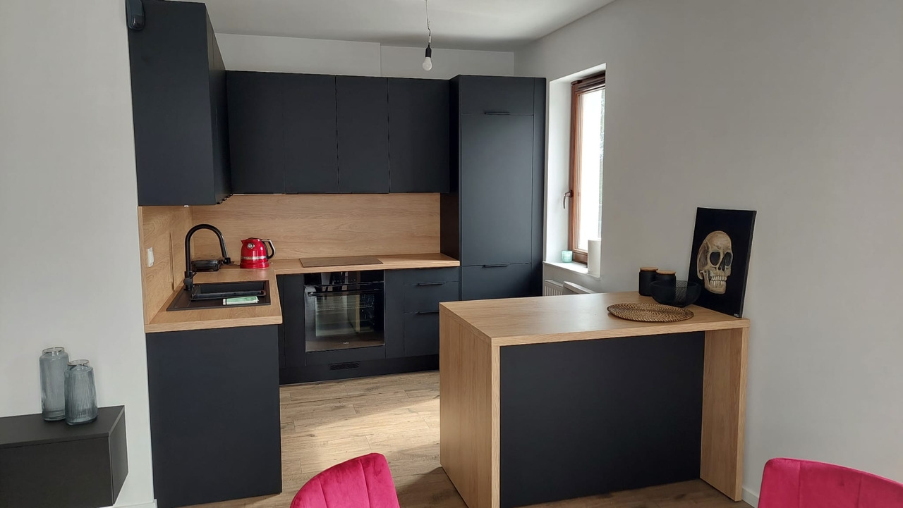 Nowoczesna kuchnia z czarnymi szafkami i blatem z jasnego drewna, uzupełniona czerwonym czajnikiem i obrazem czaszki na blacie.