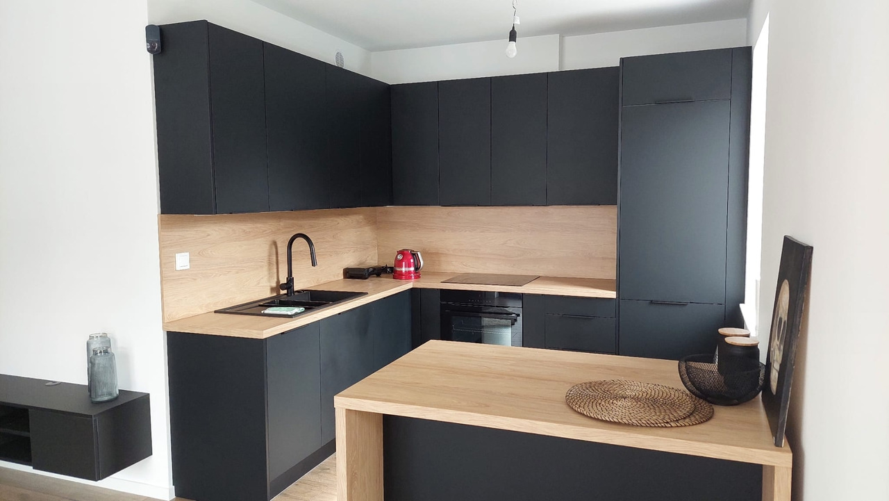 Nowoczesna kuchnia z czarnymi szafkami i blatem drewnopodobnym, oświetlona wiszącą lampą, z widocznym czarnym zlewem i czerwoną czajnikiem elektrycznym.