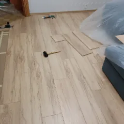 Układanie paneli laminowanych w pokoju, widoczny gumowy młotek, częściowo przykryty mebel, biała ściana w tle.