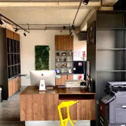 Nowoczesne biuro z drewnianą recepcją, komputerem iMac, ścianą z zieleni, półkami i żółtym krzesłem trójnożnym, oświetlone reflektorami na szynoprzewodzie.