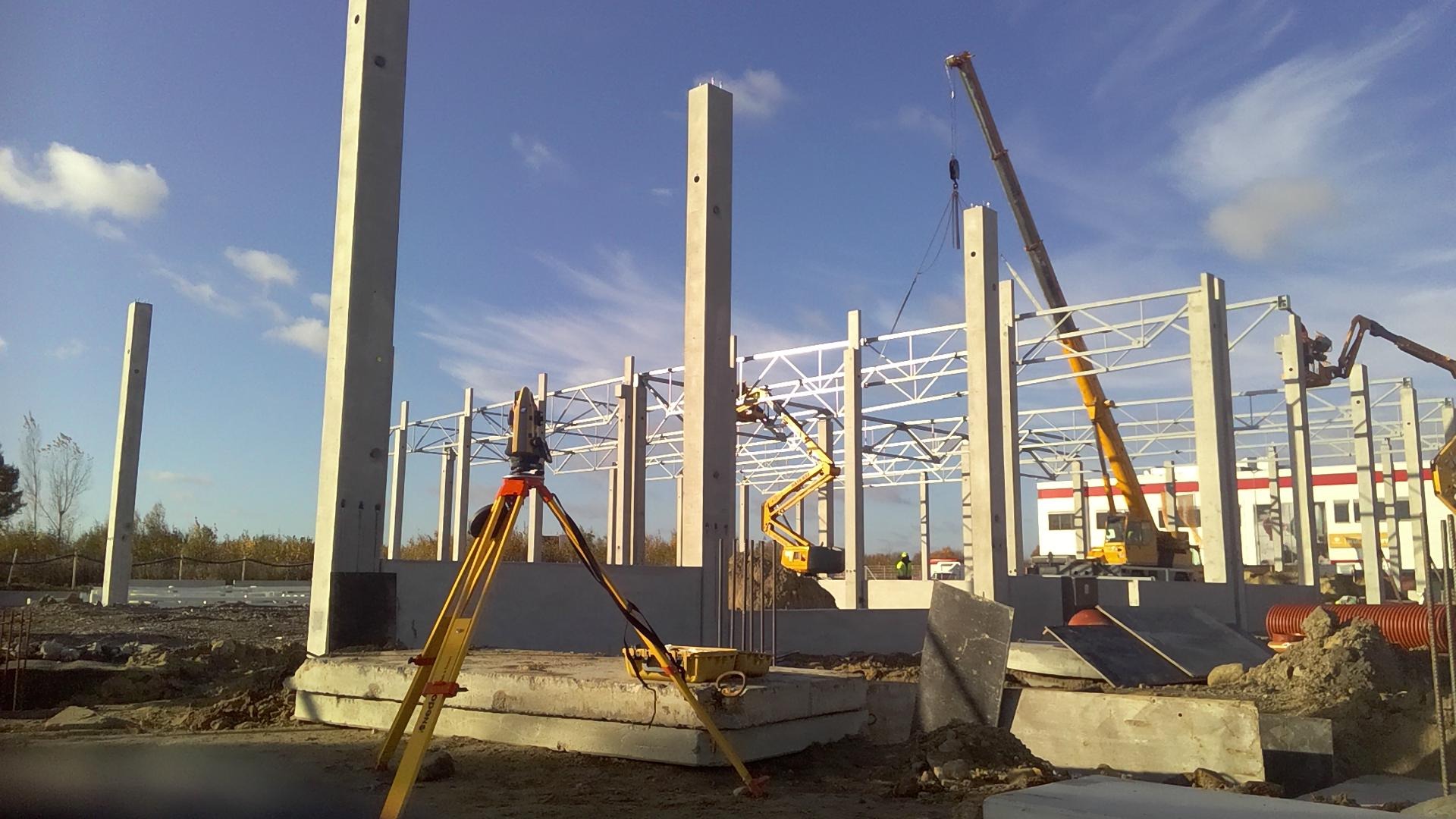 Prace budowlane w toku: widoczne betonowe słupy, stalowa konstrukcja hali, żuraw i niwelator na statywie.