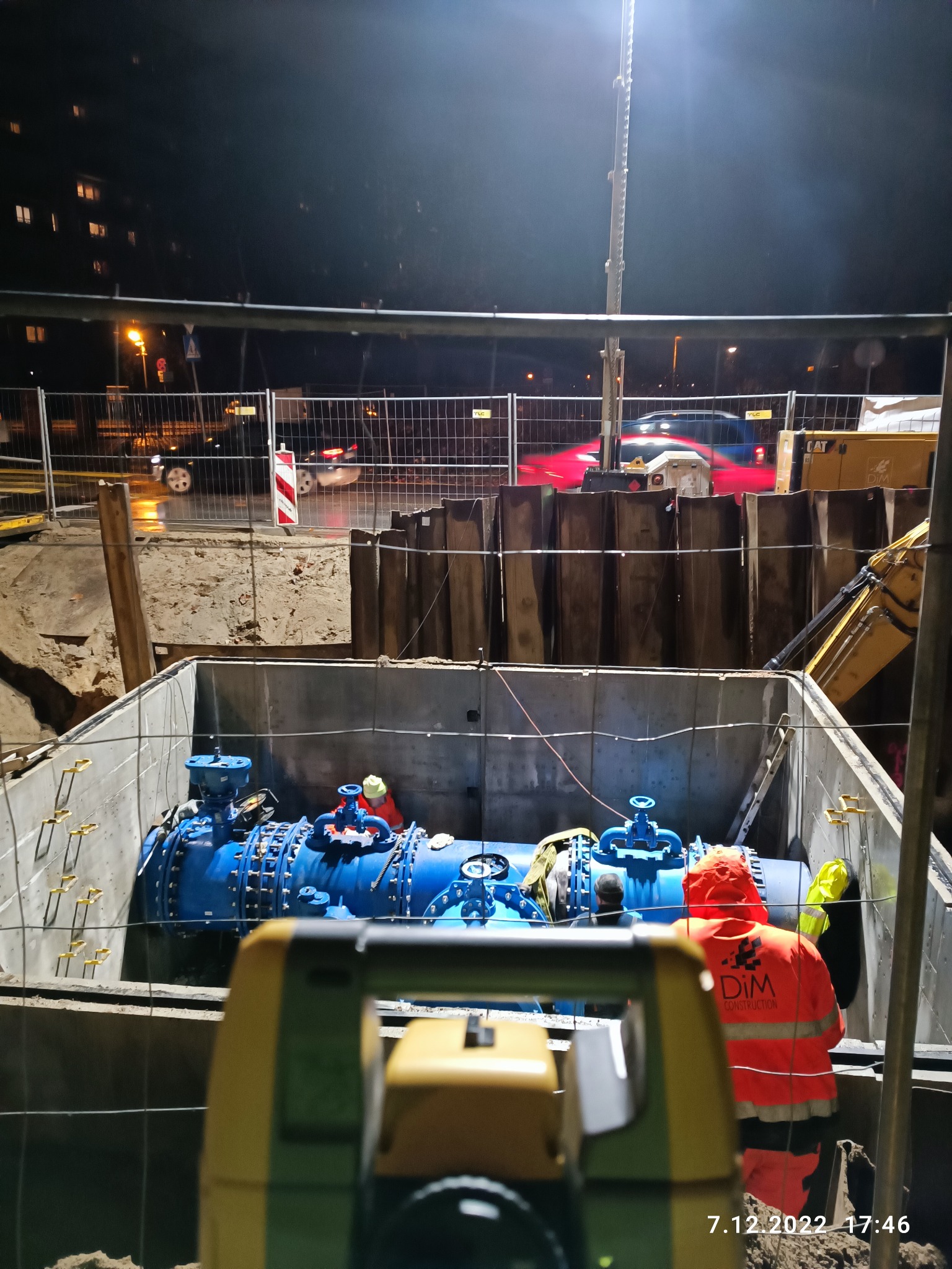 Prace instalacyjne rurociągu w wykopie zabezpieczonym szalunkiem, widoczne elementy pomiarowe w pierwszym planie i pracownicy w odblaskowych ubraniach, nocna sceneria z oświetleniem budowlanym.