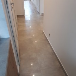 Usługi ogólnobudowlane Wojciech Zgórski - Beżowa, błyszcząca podłoga w korytarzu z widoczną listwą przypodłogową i fragmentem szafki z drewnianym frontem.