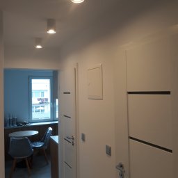 Usługi ogólnobudowlane Wojciech Zgórski - Nowoczesny korytarz z białymi ścianami i drzwiami z poziomymi wstawkami, oświetlony trzema okrągłymi lampami sufitowymi. W tle widoczny fragment kuchni z oknem, stolikiem i krzesłami.